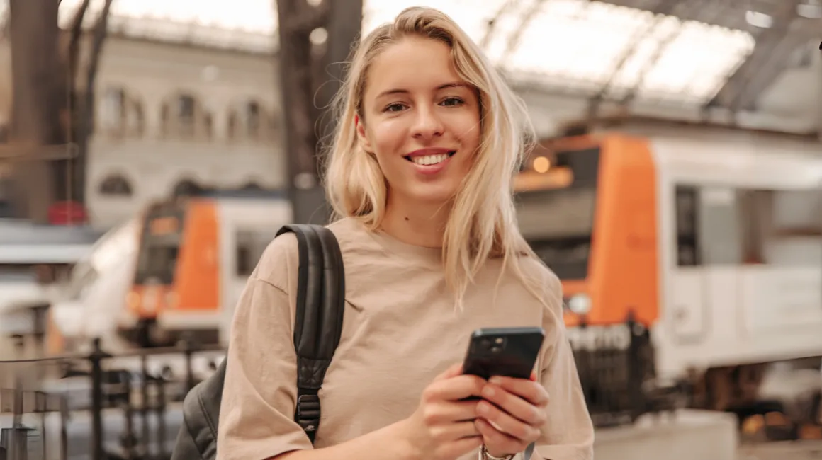 Uśmiechnięta młoda kobieta z plecakiem na dworcu kolejowym korzystająca ze smartfona